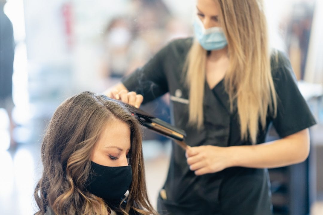 Hairdresser, protected by a mask, combing her client's hair with a hair iron in a salon